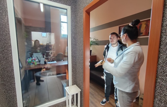 Dwie osoby w korytarzu budynku, rozmawiające przez przeszklone drzwi z osobą znajdującą się wewnątrz biura.