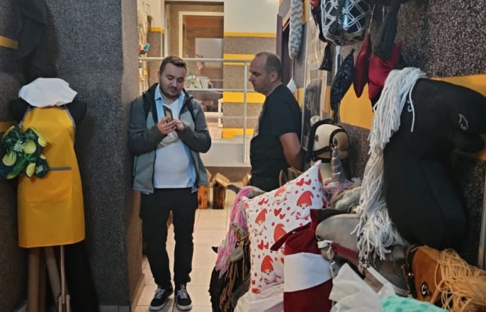 Dwóch mężczyzn rozmawiających w korytarzu budynku, otoczeni różnorodnymi ozdobami i rękodziełem na półkach i ścianach.